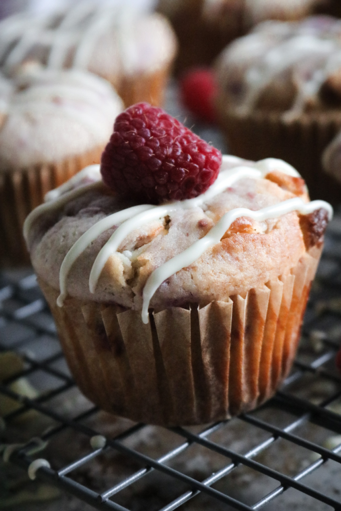 close up view of a moist white chocolate raspberry muffin with a white chocolate drizzle and fresh berry on top.