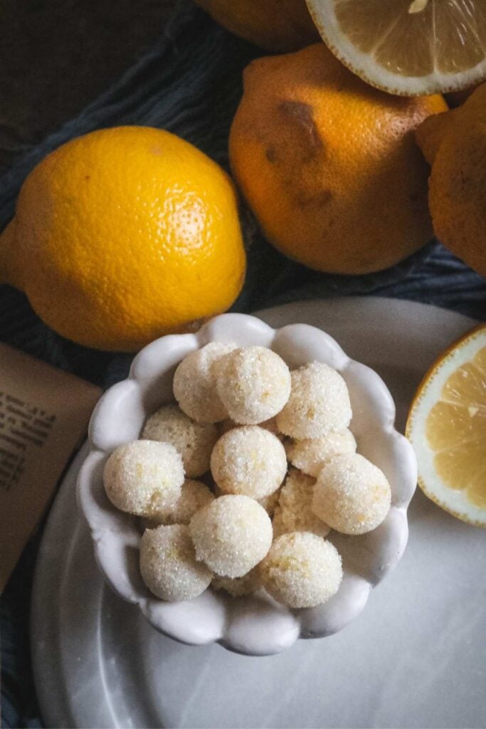 a white bowl filled with homemade lemon sugar cubes on a marble surface next to fresh lemons.