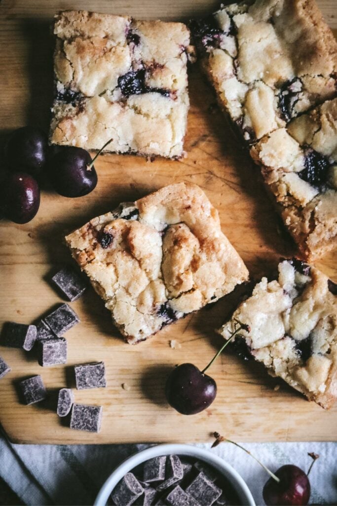 overhead view of sliced cherry blondies on a wooden cutting board next to fresh cherries and chocolate chunks.