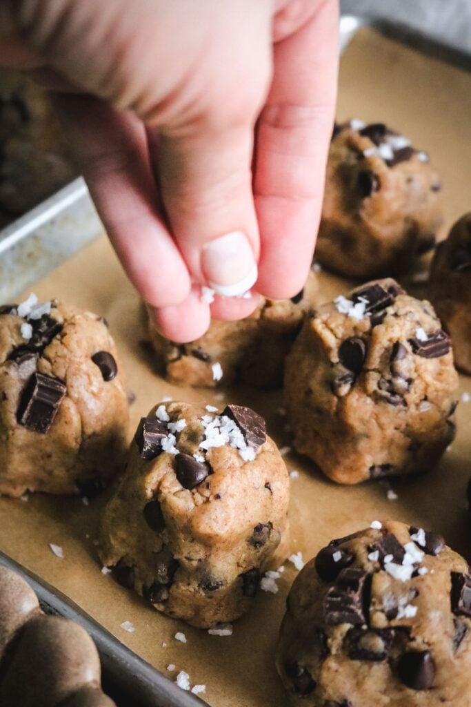 fingers sprinkling flaky sea salt over top of dough balls of homemade brown butter chocolate chip cookie dough.