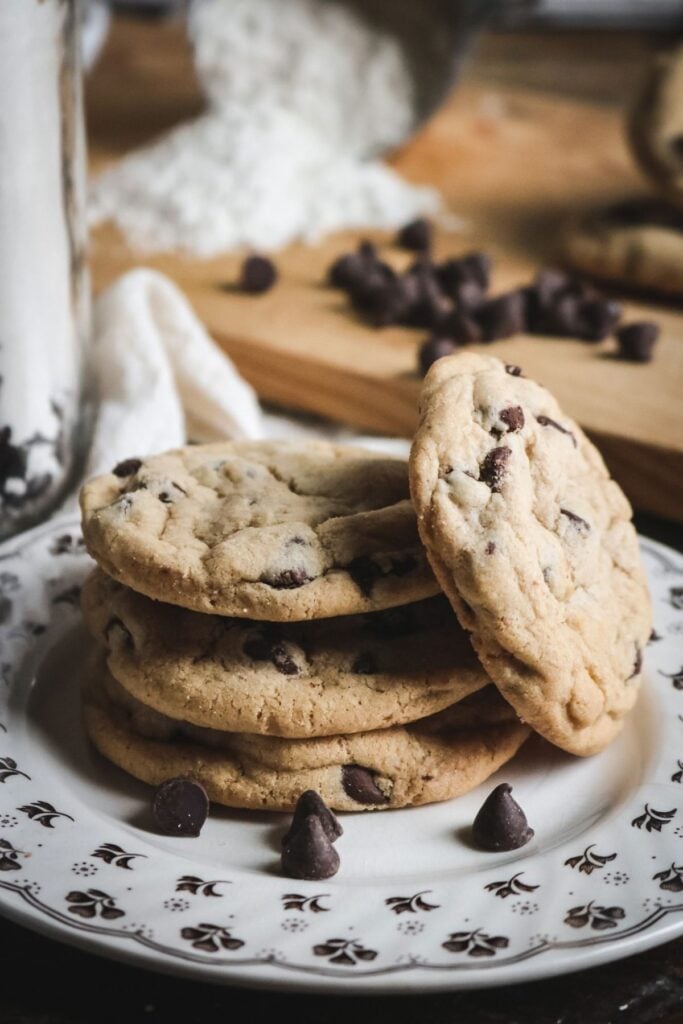 a side view of a stack of chewy chocolate chip cookies on a white and brown plate with an assortment of baking ingredients in the background.