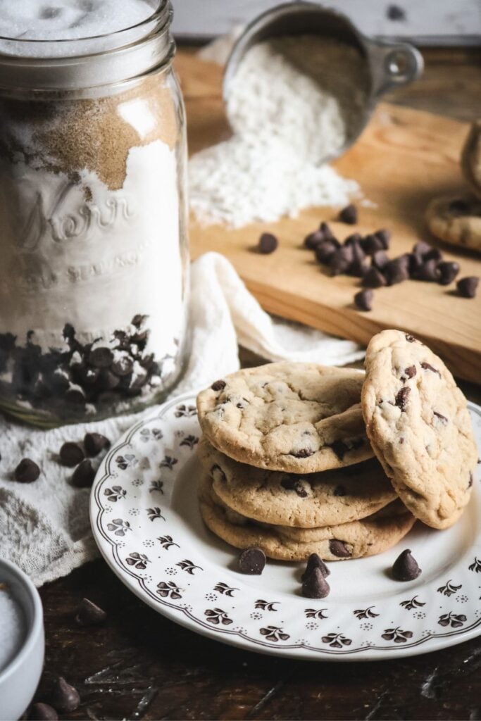 a mason jar with homemade chocolate chip cookie dry mix behind a plate of freshly baked cookies sprinkled with chocolate chips.