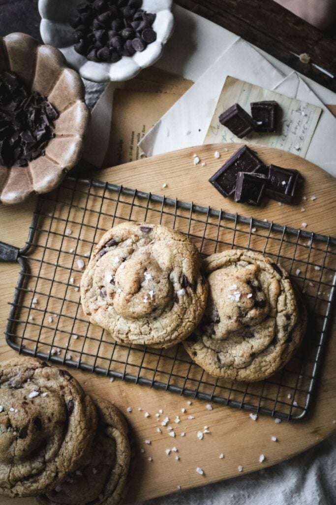 overhead view of two huge chewy brown butter chocolate chip cookies on a wire rack over a wooden cutting board surrounded by chocolate chips and chunks.
