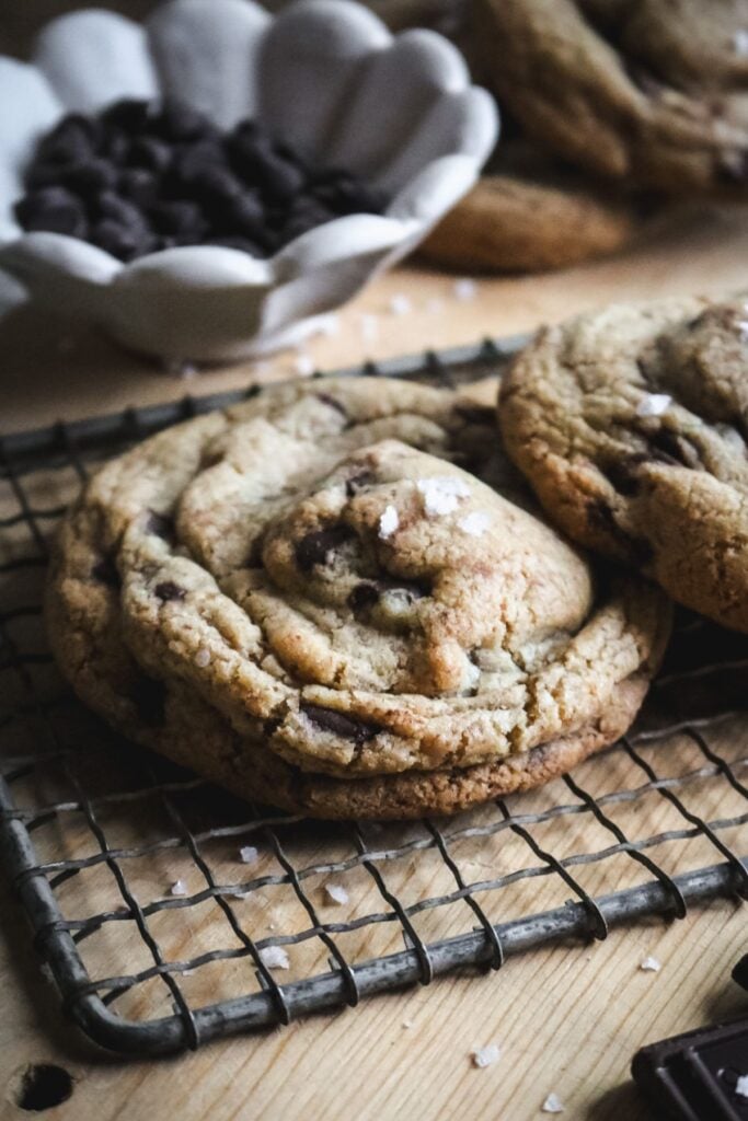 close up view of a thick, super chewy brown butter chocolate chip cookie on a wire rack in front of a bowl of chocolate chips.