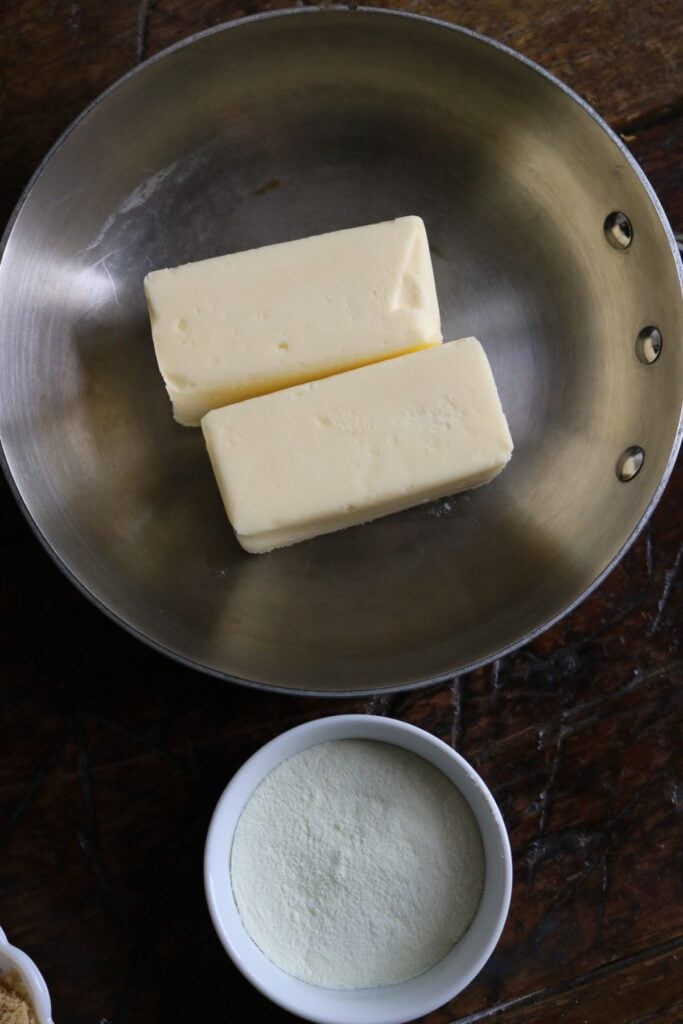 overhead view of a stainless steel frying pan with butter for browning next to a bowl of dry milk powder.