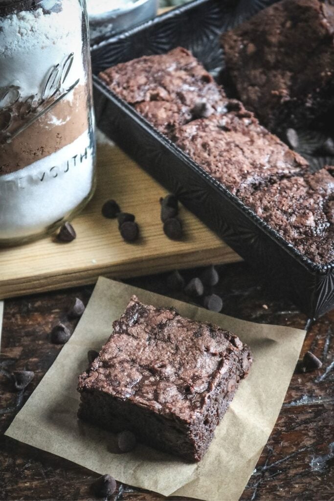 a thick square of homemade brownie next to a tray of brownies and a jar of homemade brownie mix on a wooden cutting board.
