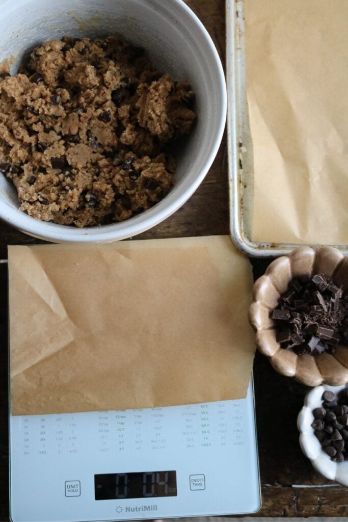overhead view of a bowl of chocolate chip brown butter cookie dough next to a digital scale and bowls of chocolate chips for shaping.