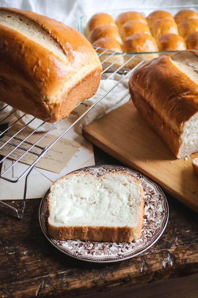 slice of soft homemade white bread on a brown transferware plate and slathered with butter with extra loaves cooling in the background.