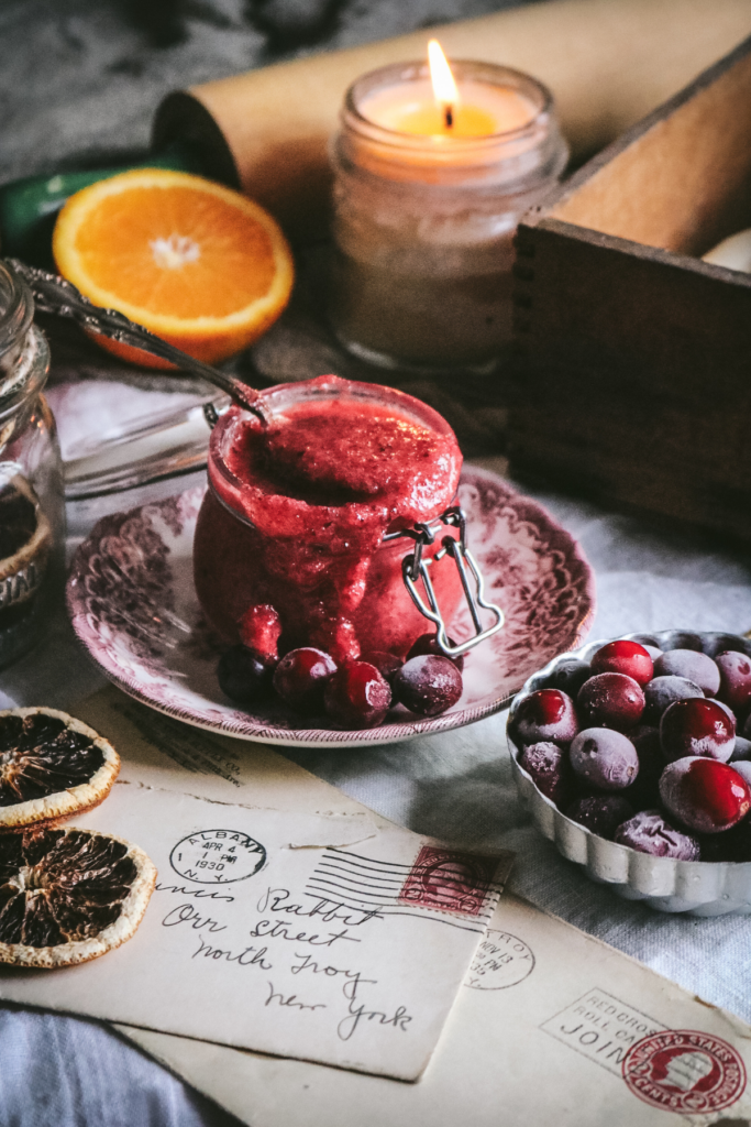 side view of a jar of sugar body scrub made with cranberries and orange zest next to a candle, frozen berries, and half an orange.