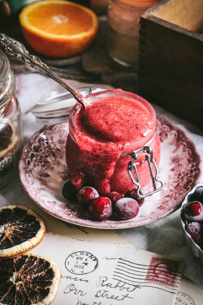 spoon scooping homemade cranberry orange body scrub next to frozen cranberries and fresh orange.