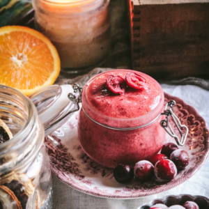 side view of a glass jar with a pink cranberry sugar scrub next to fresh orange and a candle.