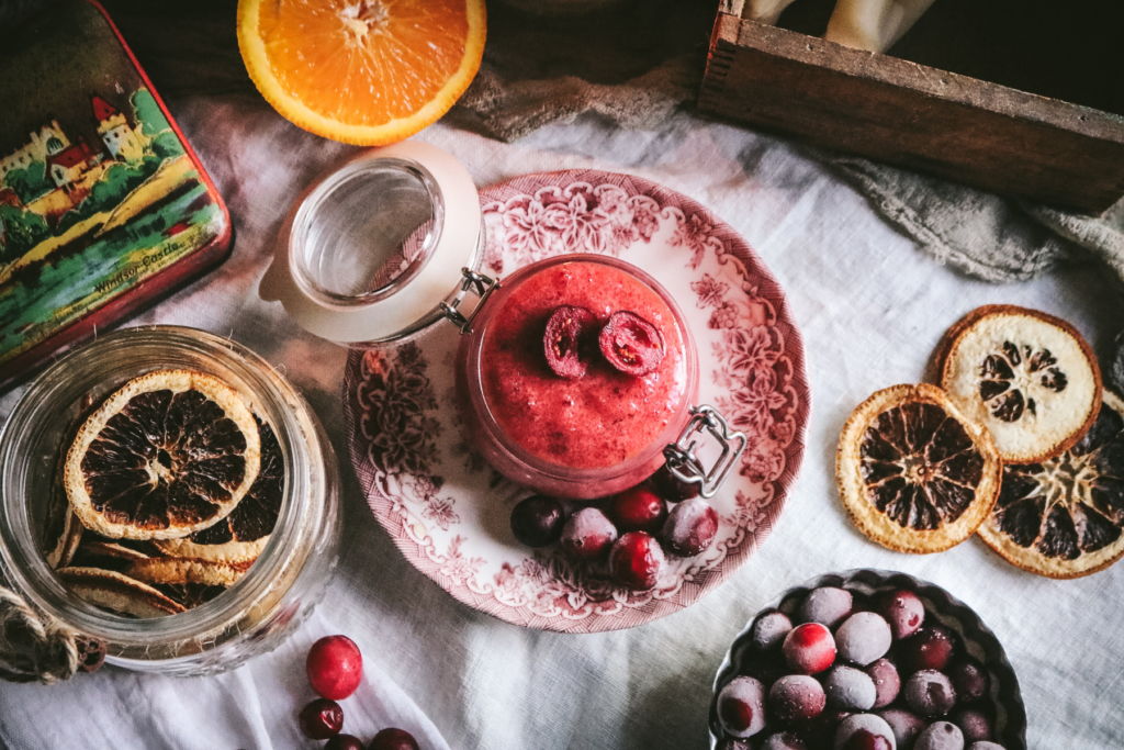 overhead view of a glass jar with homemade cranberry body scrub next to dried oranges and frozen cranberries.