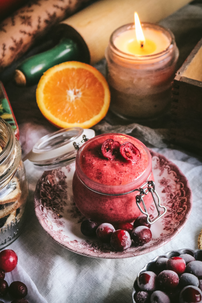 jar of homemade sugar scrub with fresh cranberries and orange zest next to a beeswax candle.