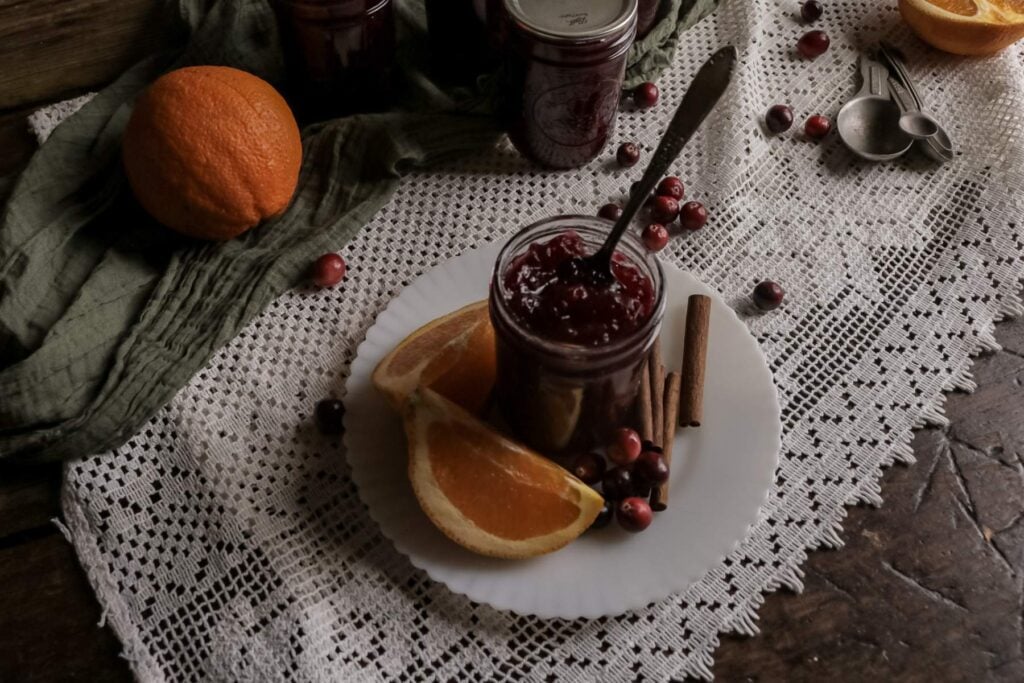 half pint mason jar with homemade cranberry sauce next to fresh oranges, cranberries, and cinnamon sticks.