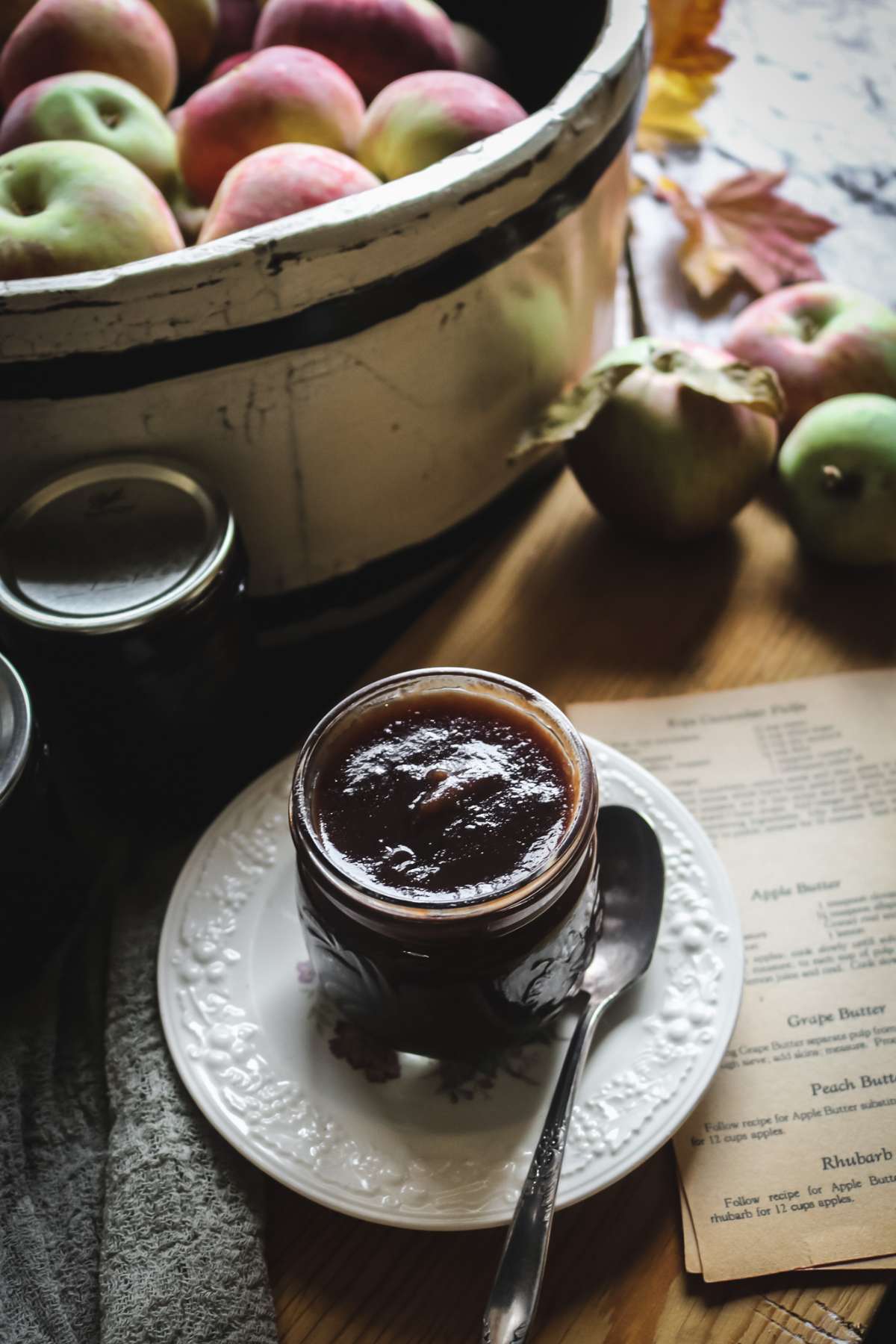 jar of apple butter on a white plate next to a barrel of fresh apples.