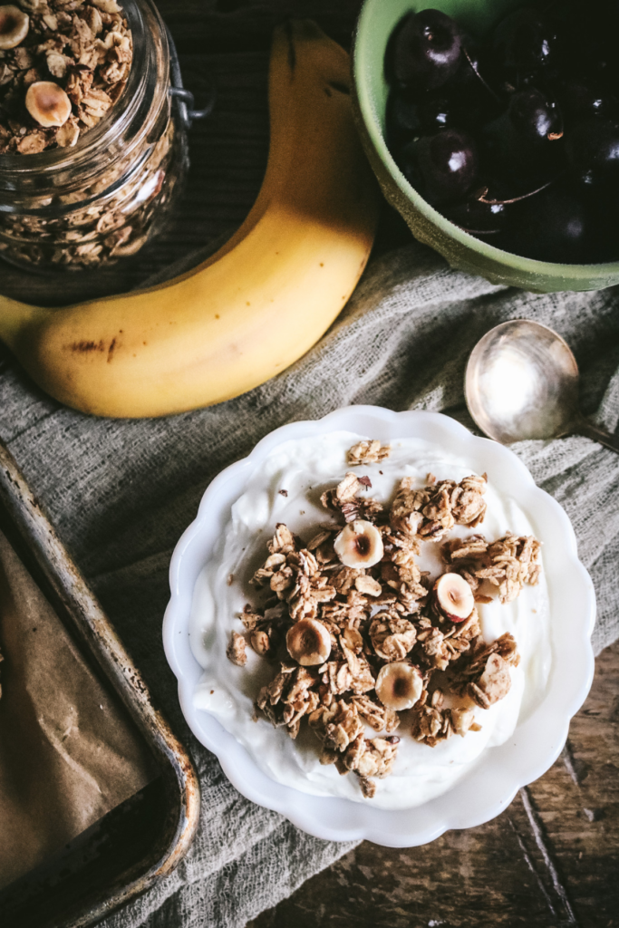 overhead view of a white dish with yogurt and hazelnut granola next to a banana and cherries.