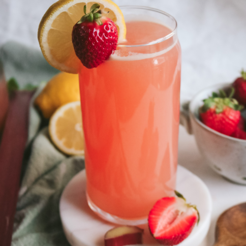 side view of a clear glass with homemade strawberry rhubarb lemonade served with fresh strawberry and lemon slices.