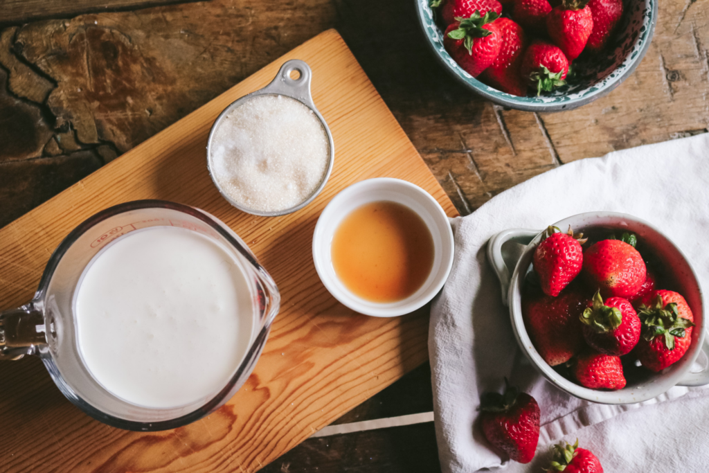 overhead view of heavy cream, sugar, strawberry extract, and fresh strawberries on a wooden table.