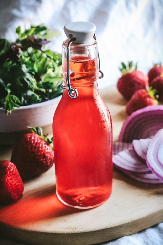 glass bottle with a reddish pink strawberry infused vinegar in front of salad ingredients.