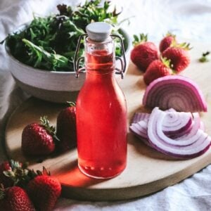 close up of a glass bottle with strawberry infused rice vinegar next to fresh berries, red onion, and salad greens.