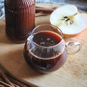 close up view of a glass creamer with homemade boiled apple cider syrup next to a fresh apple.