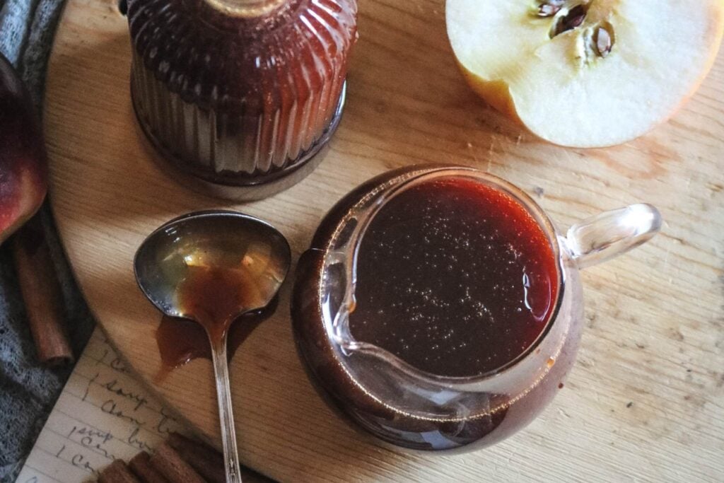 overhead view of boiled cider syrup in a glass container next to a spoon and a halved apple.
