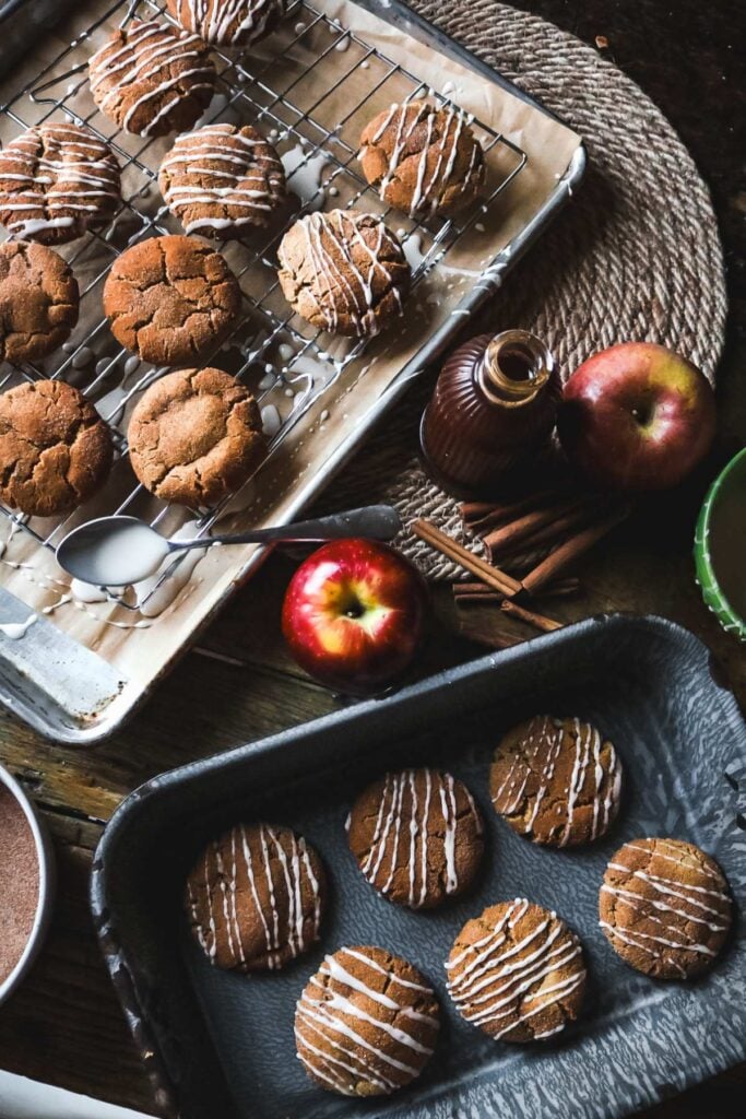 decorating apple cider cookies with apple glaze next to homemade boiled apple cider reduction and fresh apples.