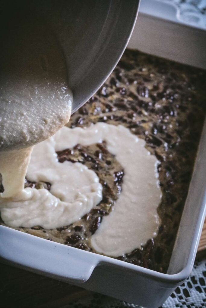 pouring cobbler batter over pecan pie filling in a ceramic baking dish.