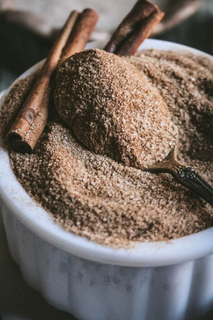 close up view of a spoonful of cinnamon sugar in a white baking dish with cinnamon sticks sticking out.