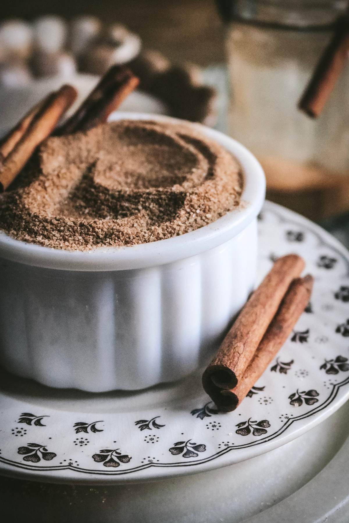 white milk glass container with homemade cinnamon sugar on a brown transferware plate next to cinnamon sticks.