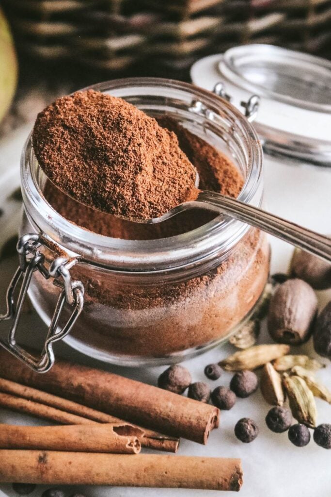 a vintage spoon heaping with homemade apple pie spice next to lots of dried herbs like cinnamon sticks, allspice berries, nutmeg, and cardamom pods.