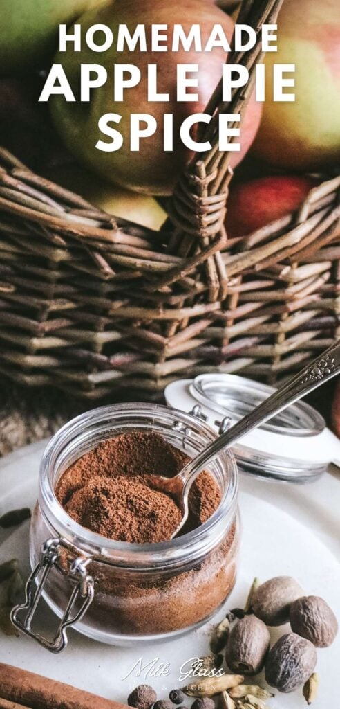 pin with an image of a homemade apple pie spice in a glass jar surrounded by dried spices and a basket of fresh apples.