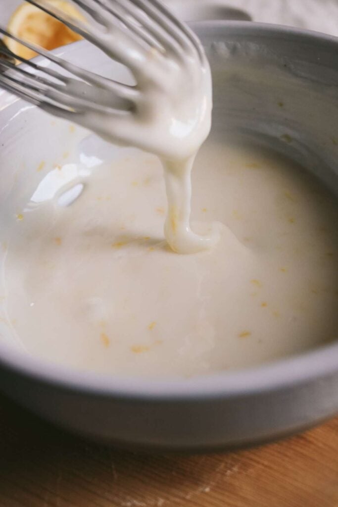 creamy lemon glaze with flecks of lemon zest in a bowl with a whisk dripping some down.