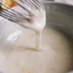 a whisk dripping homemade lemon glaze into a white bowl.