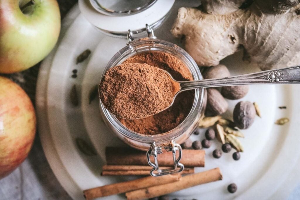 overhead view of a vintage spoon in a jar of homemade apple pie spice with fresh ginger and dried spices around the jar.