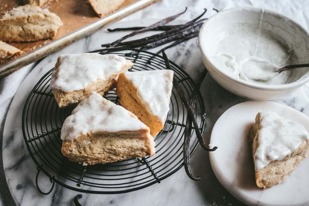 side view of vanilla bean scones cooling on a wire rack next to a bowl of vanilla bean glaze and lots of vanilla beans.