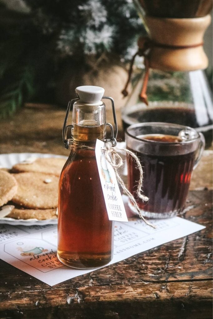 a glass bottle of almond coffee syrup in front of a plate of cookies, a chemex, mug of cookie, and a christmas tree.
