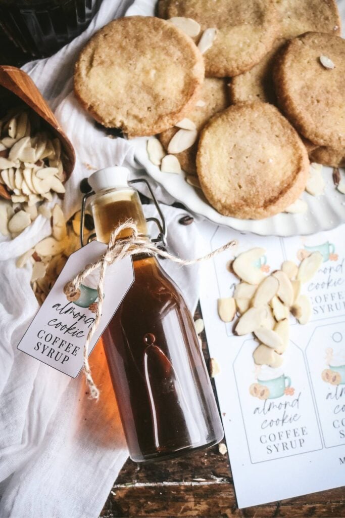 overhead view of a bottle of homemade almond coffee syrup next to shortbread cookies, printable labels, and sliced almonds for holiday gifting.
