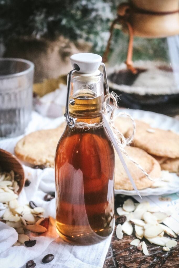 glass bottle glowing bright with an amber colored homemade almond coffee syrup in front of cookies, a mug of coffee, a chemex, and a christmas tree.