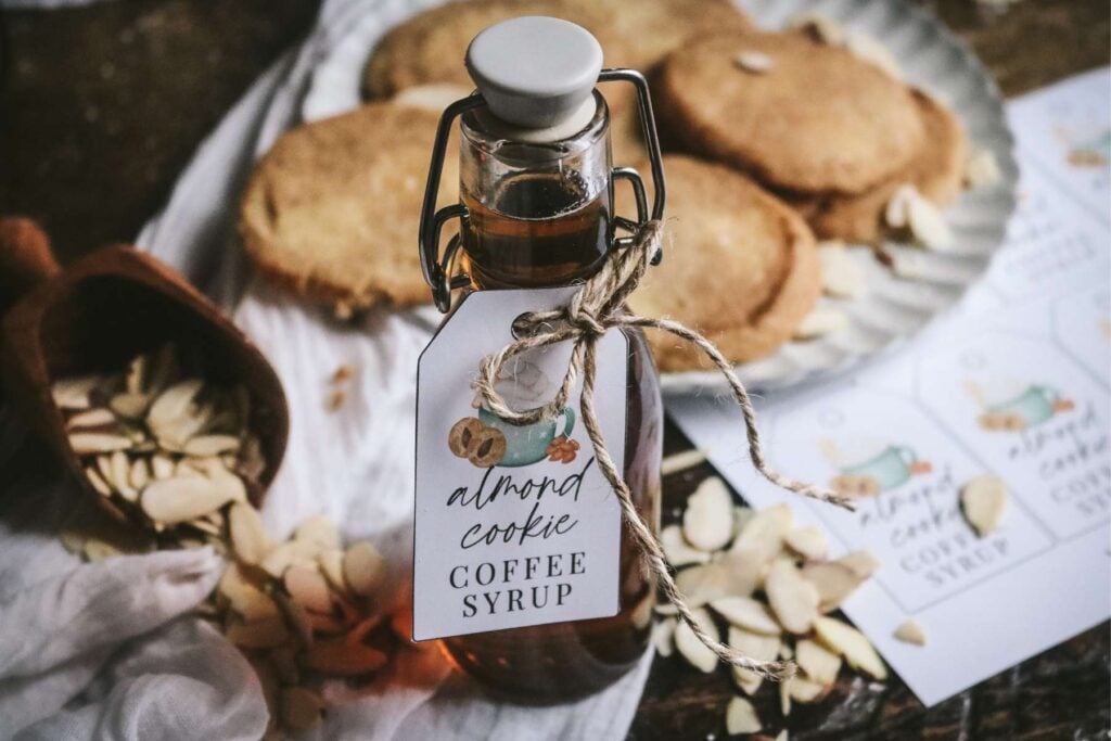 a bottle of homemade almond coffee syrup in front of shortbread cookies next to a scoop of almonds and printed gift tags.