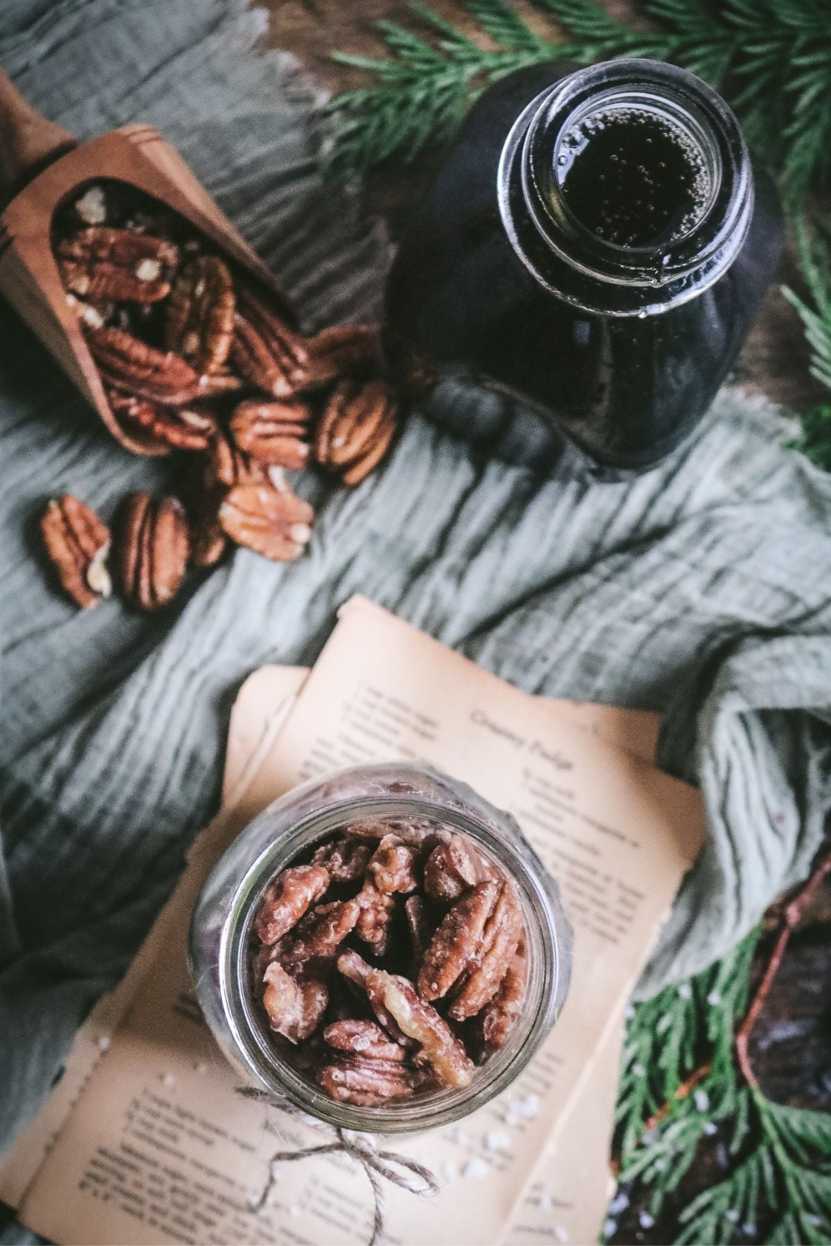 overhead view of a jar of maple pecans on antique cookbook pages next to cedar greens, maple syrup, and fresh pecans.