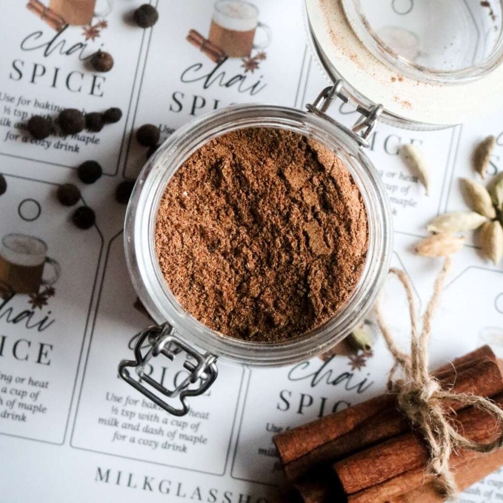overhead view of a spice jar of homemade chai mix over printable labels next to cinnamon sticks, cardamom, and allspice berries.