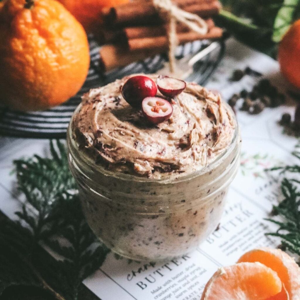 close up view of a glass jar with homemade cranberry compound butter with sliced cranberries on top, cedar boughs, fresh oranges, and cinnamon sticks on top of printed labels for gifting.