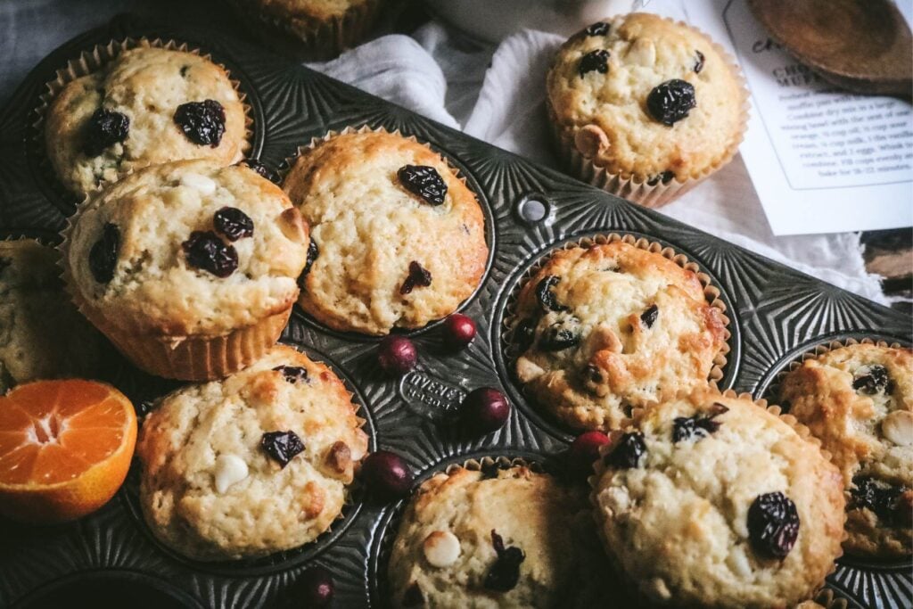 overhead view of cranberry white chocolate muffins with orange in an antique muffin tin next to fresh cranberries.