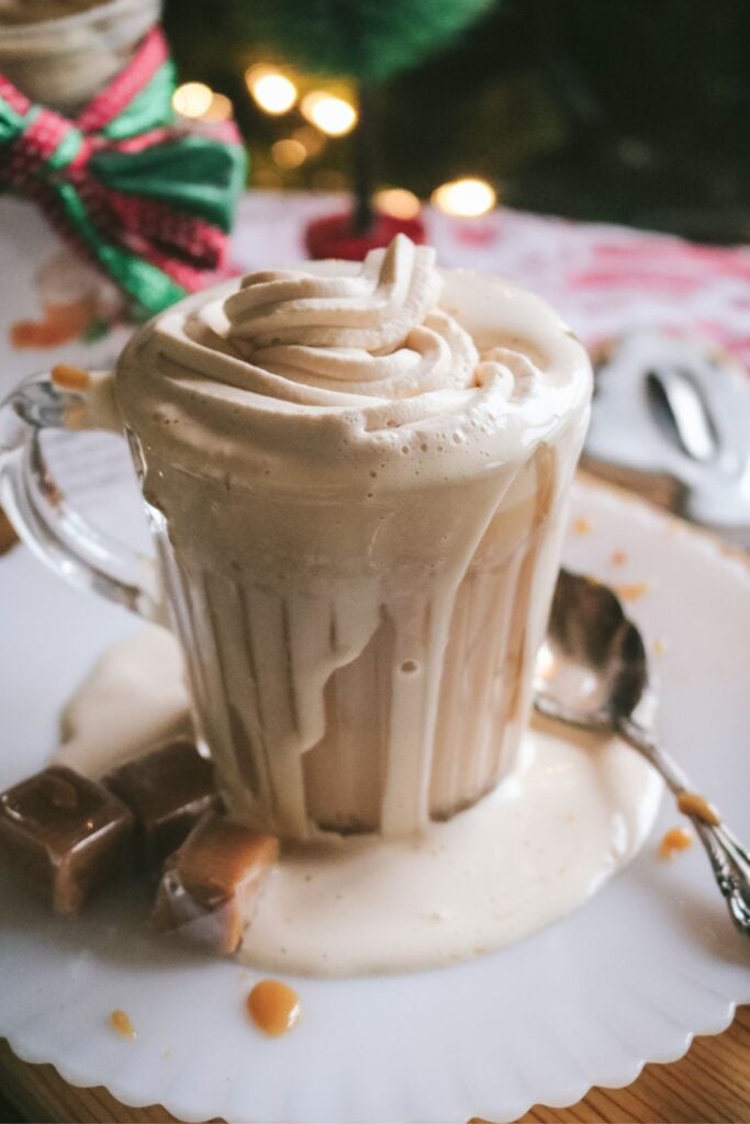 glass mug overflowing with homemade hot caramel milk made with a homemade dry mix and topped with caramel whipped cream and caramel drizzle with a christmas tree in the background.