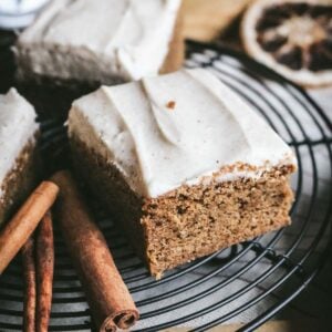 close up view of chewy gingerbread blondies with cinnamon cream cheese frosting on a wire rack next to cinnamon sticks.