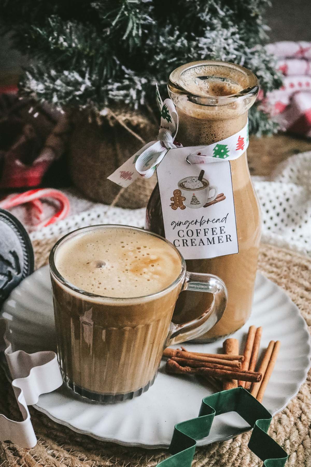 a glass mug of coffee next to a jug of from scratch gingerbread coffee with candy canes, cookie cutters, and a christmas tree in the background.