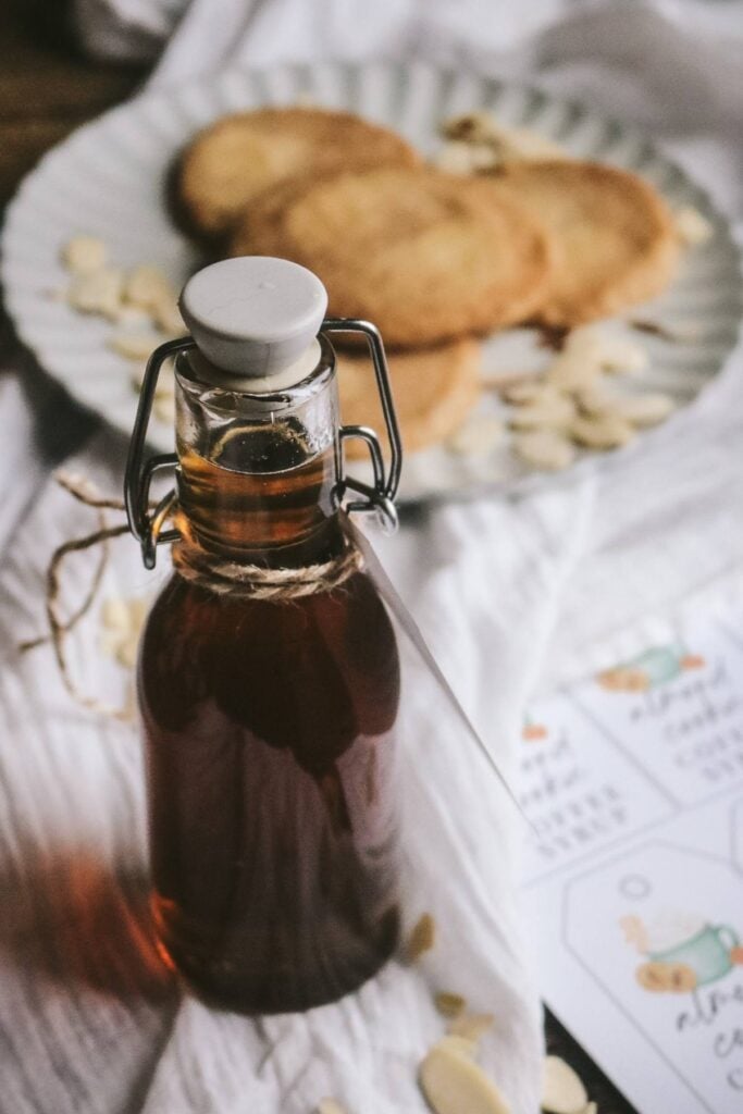 a glass bottle of amber-colored cookie-flavored almond syrup in front of a tray of cookies next to printable gift tags.