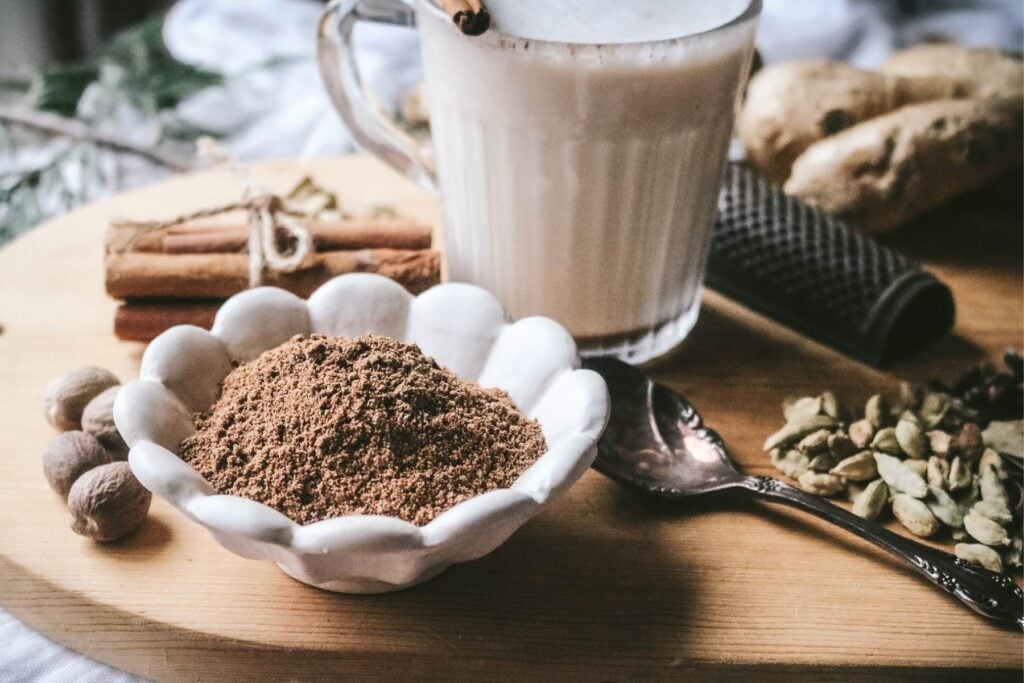 white dish next to a glass mug of creamy homemade maple chai next to lots of spices, like nutmeg, cardamom, cinnamon, and ginger.