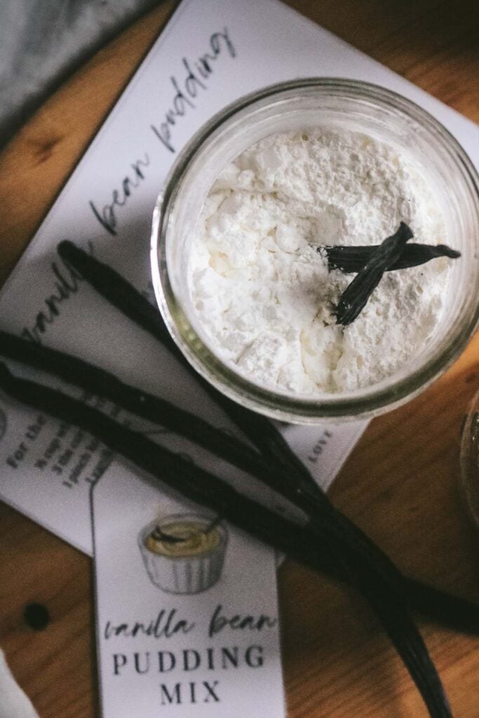 overhead view of a jar of homemade vanilla bean pudding mix including a whole vanilla bean and printable tags and a recipe card with instructions.